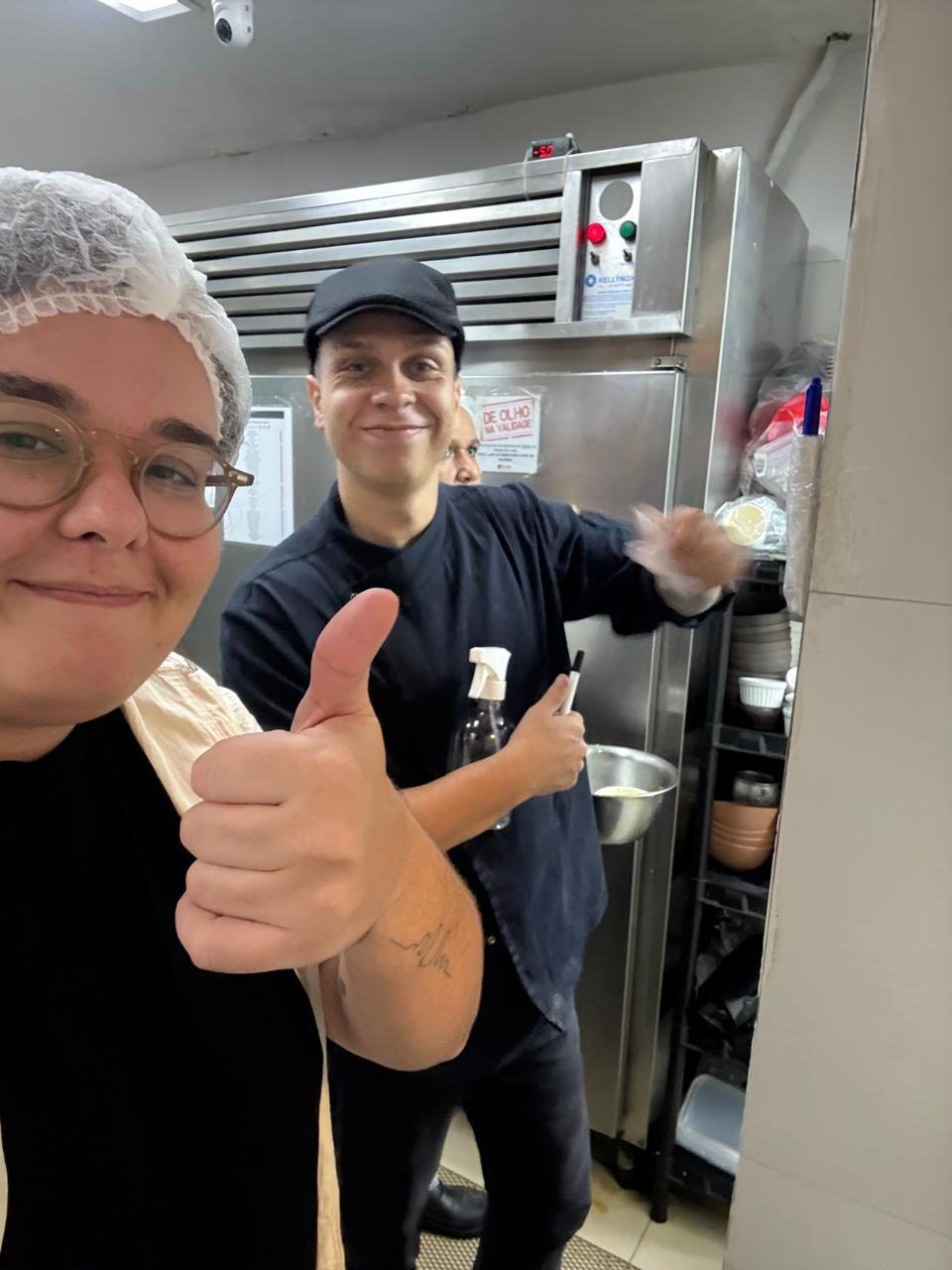 Selfie na cozinha de um restaurante: duas pessoas sorrindo, uma de touca e óculos fazendo sinal de positivo, outra de dólmã escuro e boné, na frente de uma geladeira de inox.